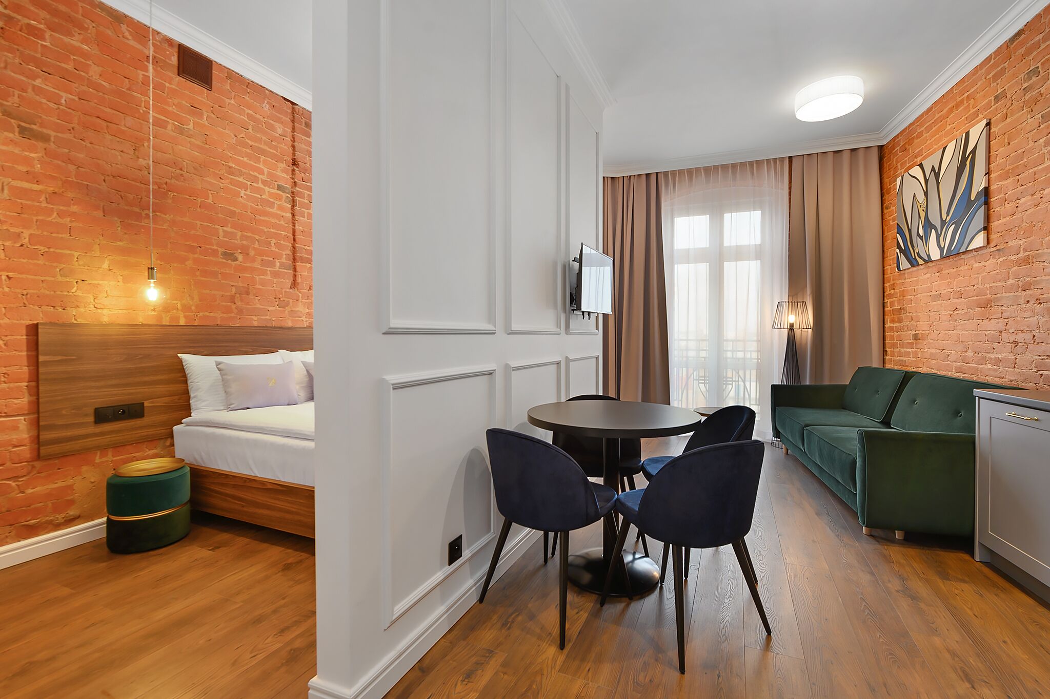 A room with a brick wall, a wooden headboard bed, a round table with chairs, a green sofa, and a TV mounted on the wall.