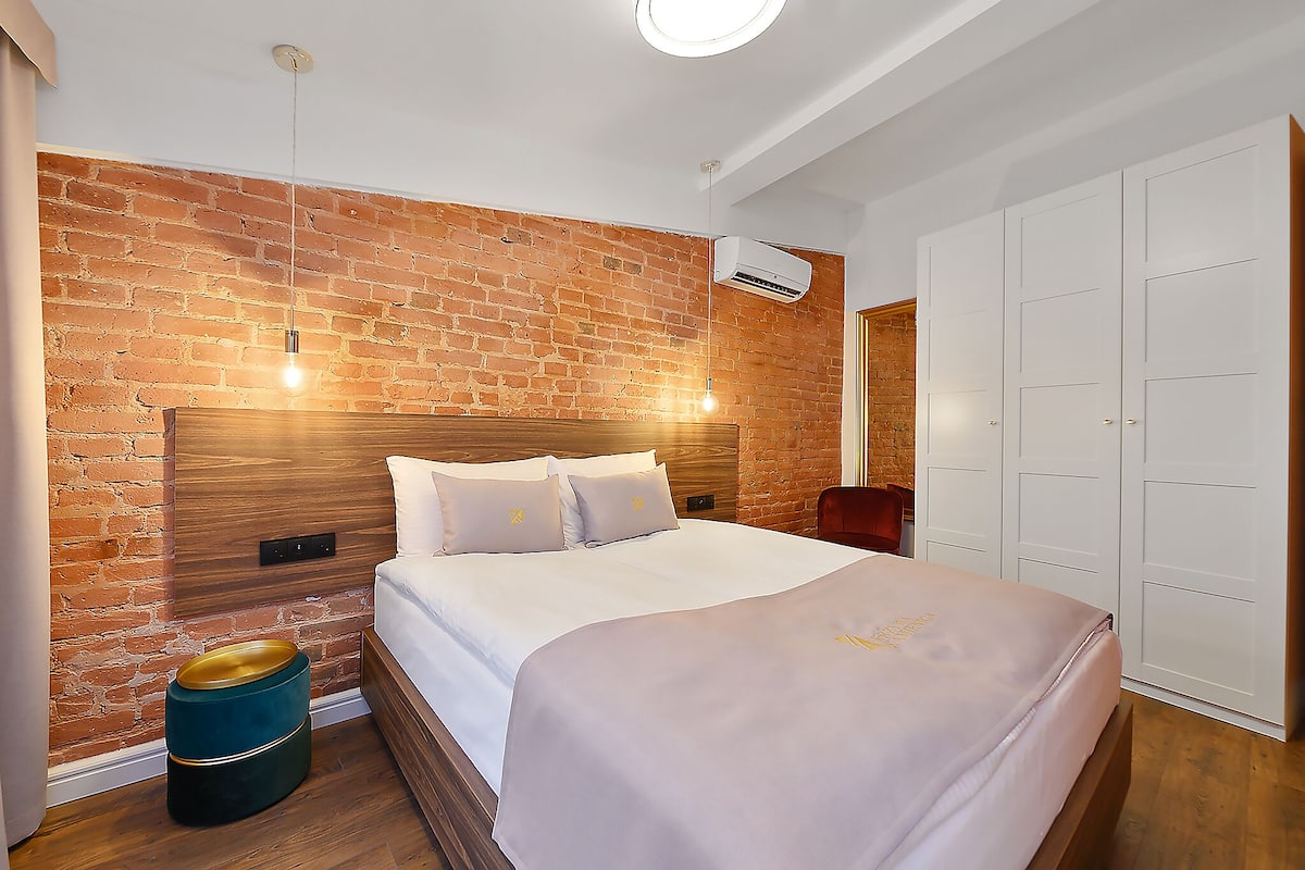 Ein Schlafzimmer mit Backsteinwand, Holz-Kopfteil, einem Bett mit weißer Bettwäsche, einem runden Mülleimer und einem weißen Kleiderschrank.