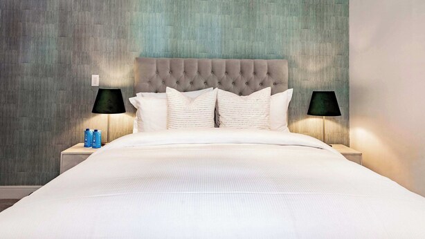 A neatly made bed with a tufted headboard, two bedside lamps, and a nightstand with a bottle and a small object.