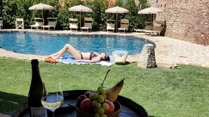 Zona de piscina con una persona recostada sobre una toalla, una botella de vino y una cesta de fruta sobre un barril.