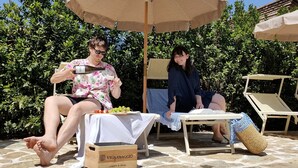 Dos personas descansando bajo un paraguas grande, con una mesa entre ellas que sostiene un cajón con la etiqueta "VIGNAMAGGIO" y una botella.