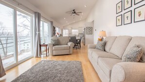 Un salon spacieux avec une grande fenêtre, un ventilateur de plafond et un coin repas à l’arrière-plan.
