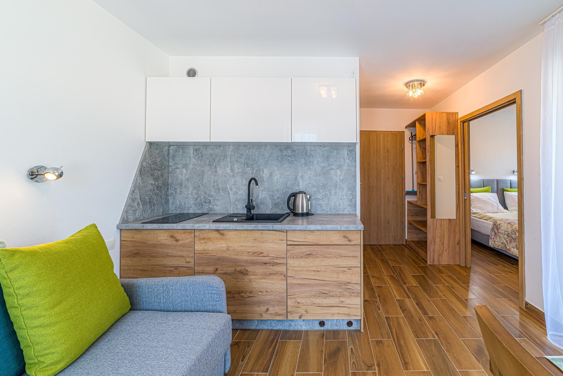 Eine kompakte Küche mit Spüle, Wasserkocher und Holzschränken. Im angrenzenden Raum befindet sich eine Couch mit einem grünen Kissen und ein Bett mit Kopfteil.