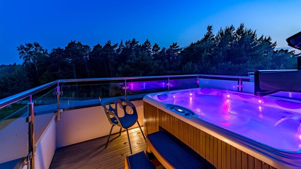 A hot tub area with a wooden deck, glass railing, and a view of a forested landscape. - Wicie Residence (Darlowo)