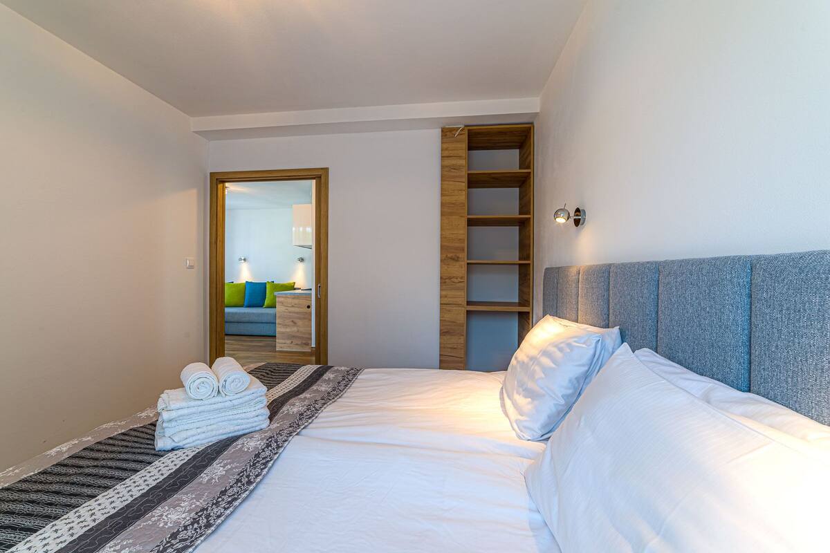 Habitación de hotel con cama, estantería de madera, baño visible a través de una puerta abierta y una pequeña pila de toallas.