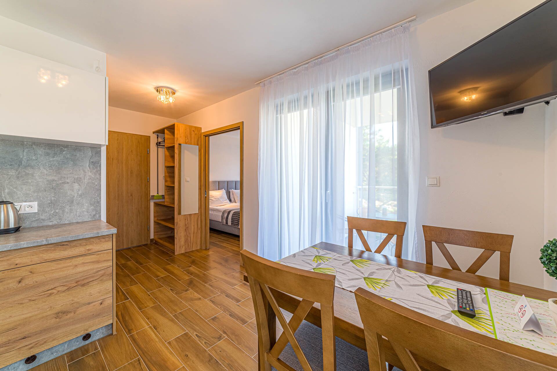 A compact living space with a kitchenette, a dining area with wooden chairs, a bed with a headboard, and a TV mounted on the wall.