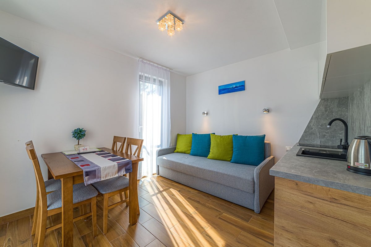 Una sala de estar moderna con un sofá gris, una mesa de comedor de madera y sillas. Hay una planta pequeña sobre la mesa, un televisor montado en la pared y una zona de cocina con un fregadero y un hervidor.