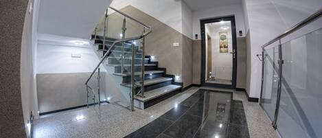 Un intérieur moderne avec un escalier en verre, un couloir menant à une porte et un sol carrelé.
