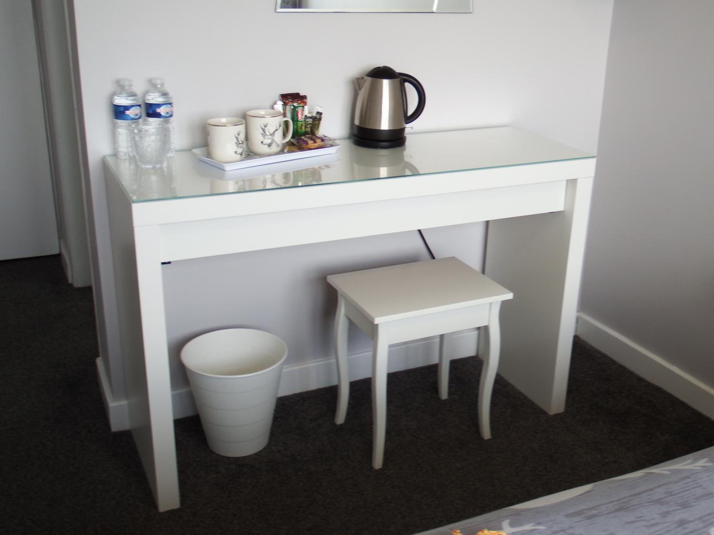 Escritorio blanco con cubierta de vidrio, cafetera de acero inoxidable, dos tazas y una bandeja con refrigerios y un cepillo de dientes.