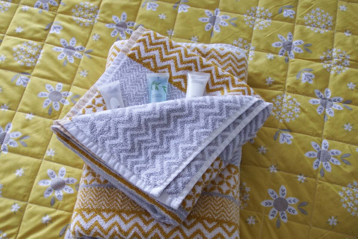 A bed with a yellow floral quilt, a folded towel, and two toiletry bottles.