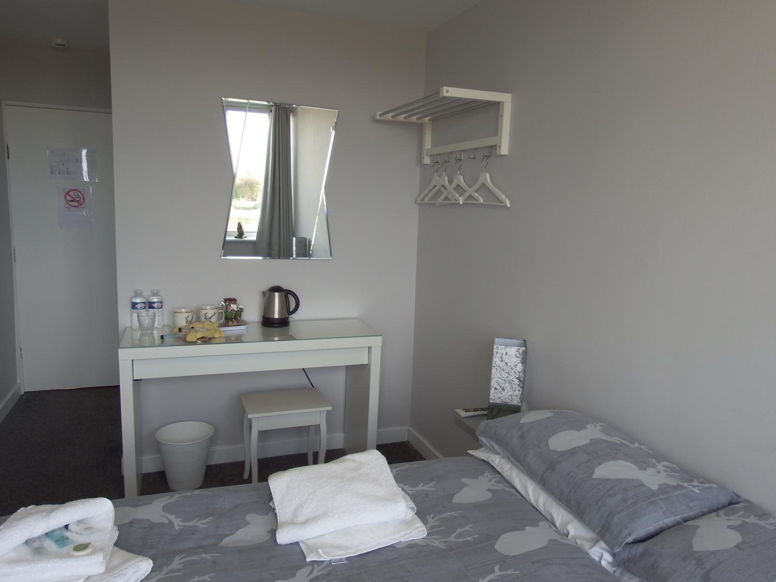 A small, clean room with a bed, a desk, and a mirror. There is a window with curtains, a towel on the bed, and a small table with a kettle and cups.