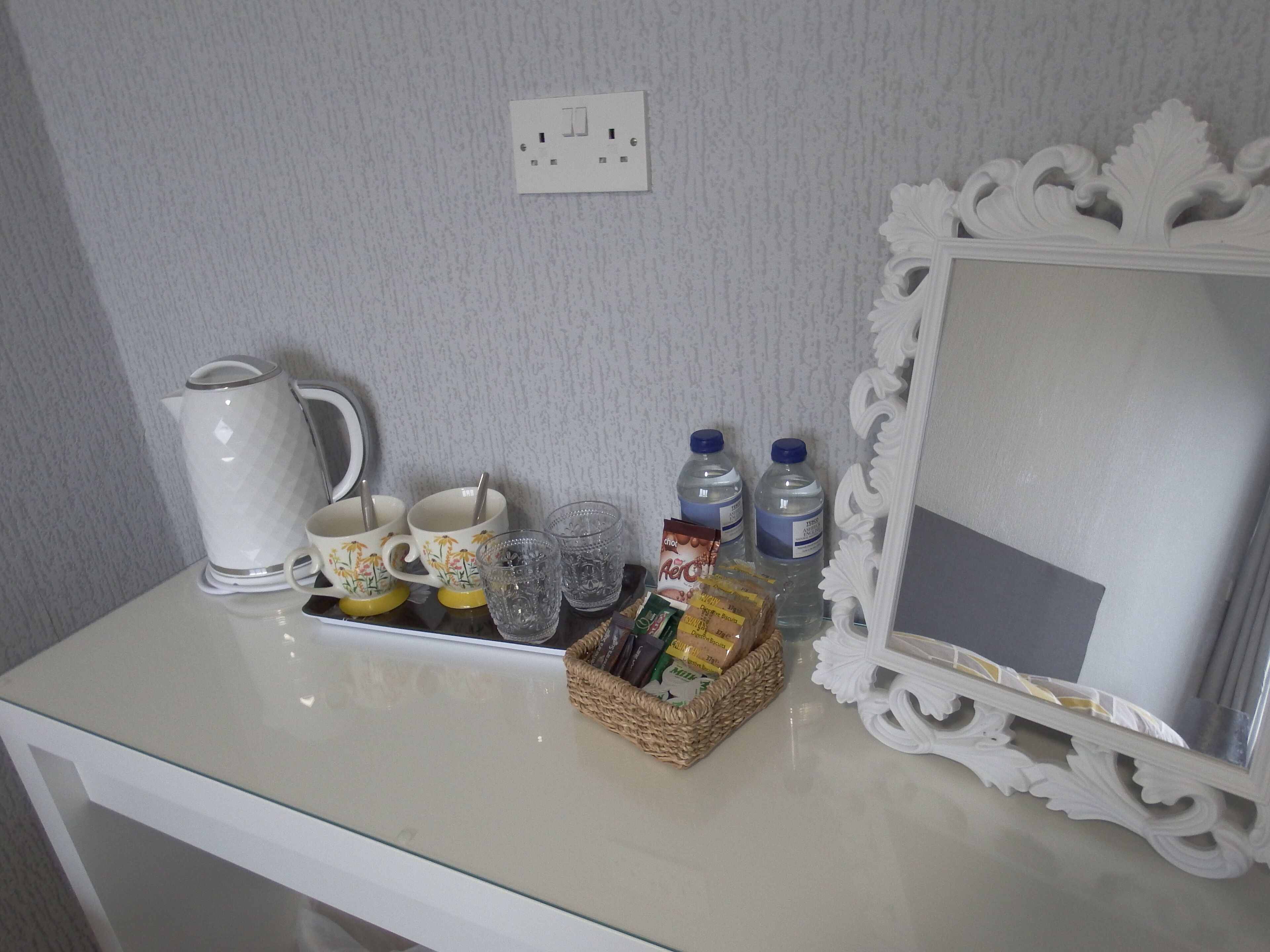 A white table with a kettle, cups, glasses, water bottles, and a basket of snacks.