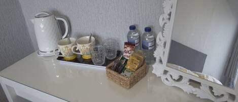 A white table with a kettle, cups, glasses, water bottles, and a basket of snacks.