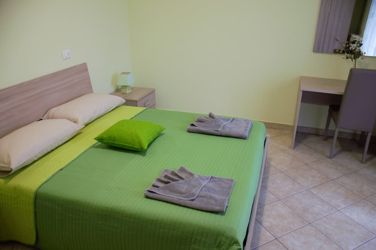 Un lit bien fait, avec une literie verte, deux oreillers et des serviettes pliées. Une petite table de chevet avec une lampe se trouve à droite, ainsi qu’une chaise.