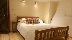 A bedroom with a wooden bed, white bedding, and a plaid pillow. There is a nightstand with a glass of wine and a lamp. The room has wooden flooring and a sloped ceiling. - Pershbrook Cottage & Garden on the River Severn. (Minsterworth)