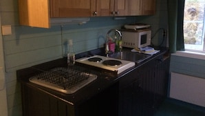 A compact kitchen with wooden cabinets, a microwave, a sink, and a window with curtains. - Lofoten Hostel (Vestvågøy)