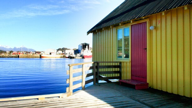A wooden dock leading to a yellow building with a red door, situated by a calm body of water with boats and distant mountains. - Lofoten Hostel (Vestvågøy)