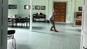 A room with a piano, dining table, chairs, and a person walking. - Lofoten Hostel (Vestvågøy)