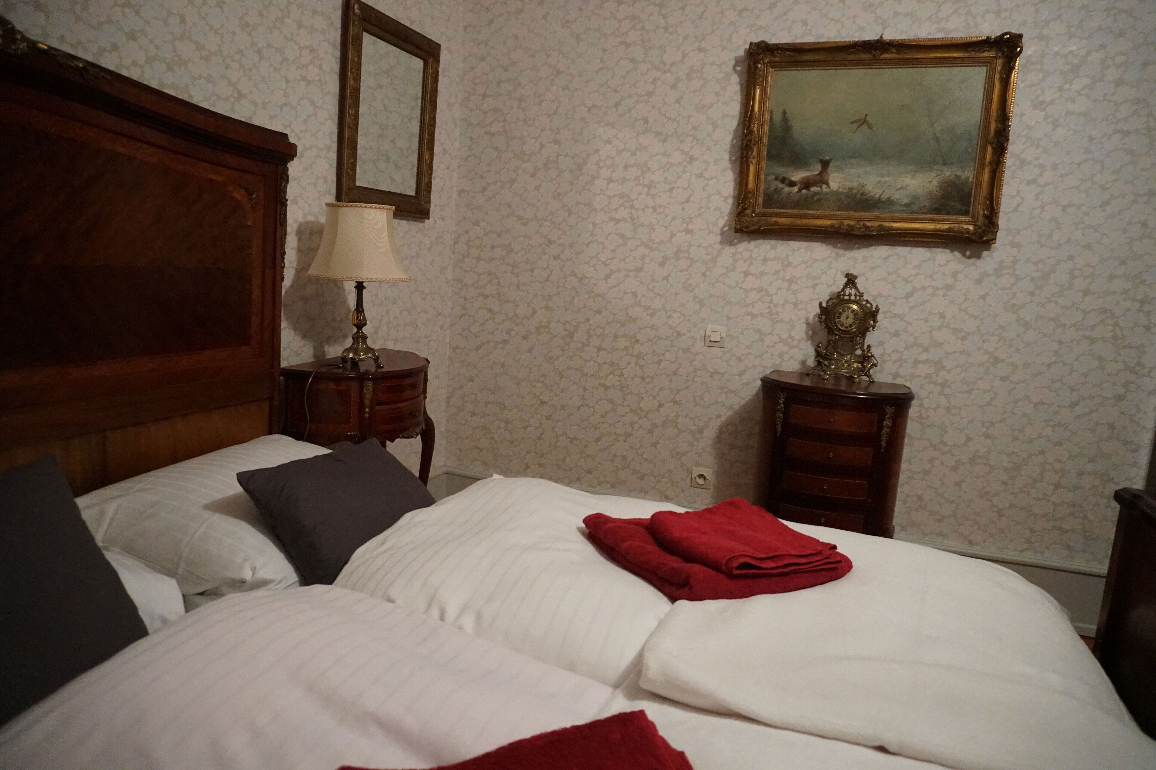 A hotel room with two beds, a wooden headboard, a nightstand with a lamp, a painting on the wall, and a small table with a clock.