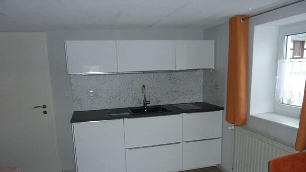 A compact kitchen with white cabinets, a black countertop, and a window with orange curtains. - Cozy apartment 5 (Zeulenroda-Triebes)