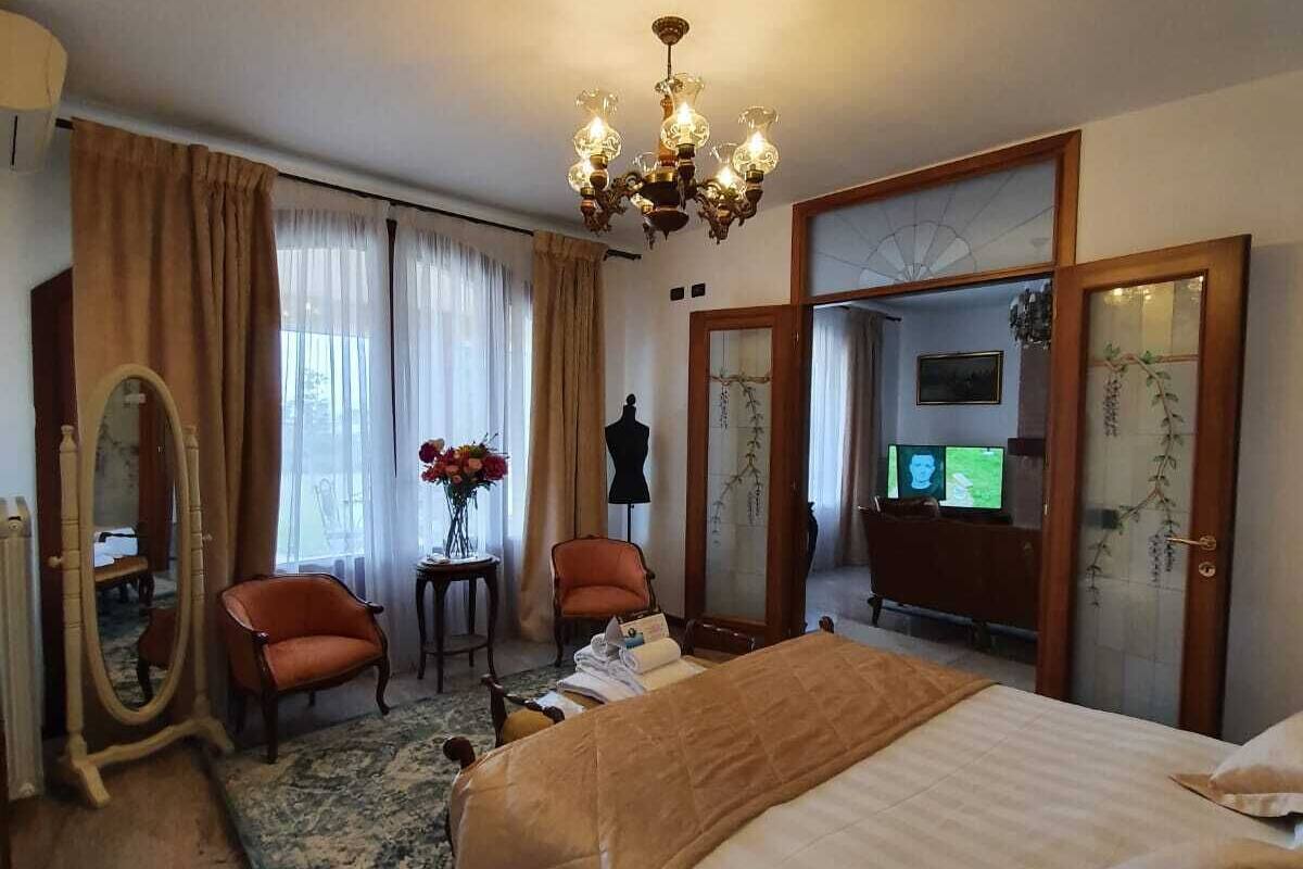 A hotel room with a large bed, a sitting area with two chairs, a vanity with a mirror, and a chandelier.