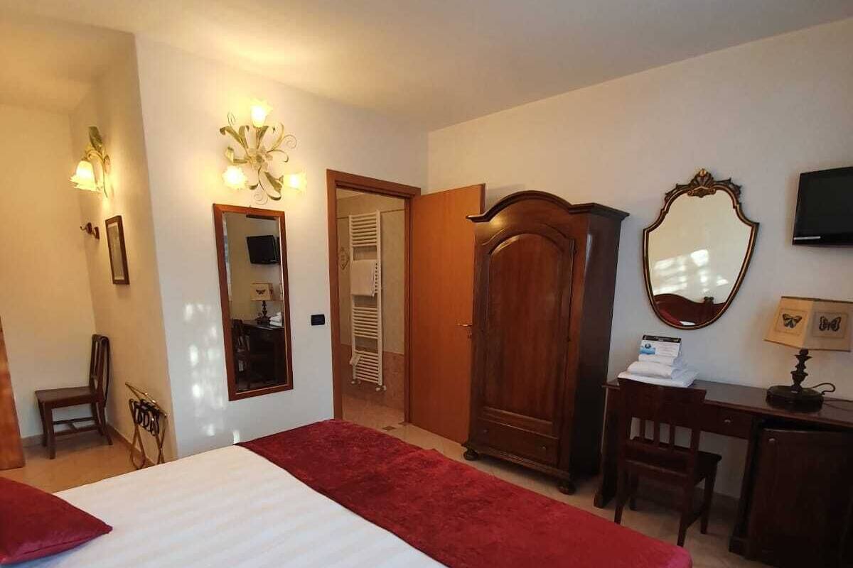 A hotel room with a large bed, a wooden wardrobe, a desk with a chair, a mirror, and a television on the wall.