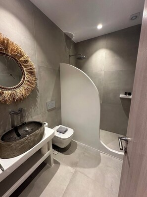 A modern bathroom with a round mirror, a stone sink, a toilet, and a shower area.