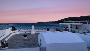Une terrasse sur le toit offrant une vue sur la mer au coucher du soleil, avec un bâtiment blanc, des plantes en pot et des sièges d’extérieur.