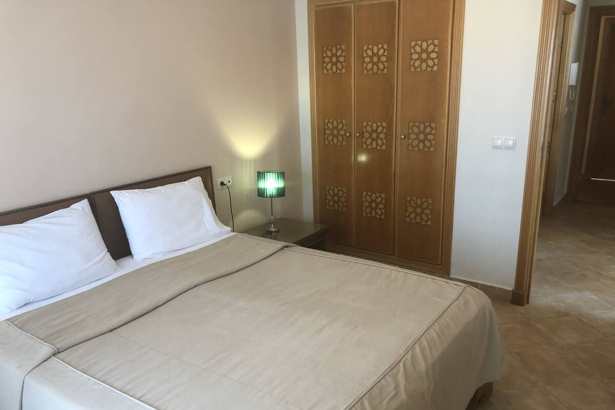 A neatly made bed with a beige bedspread and white pillows, a bedside table with a green lamp, and a wooden wardrobe with decorative panels.