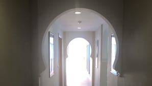 Un pasillo largo y estrecho con piso de baldosas y paredes blancas. Hay varias puertas a ambos lados, que dan a otras habitaciones.