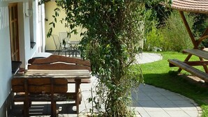 An outdoor seating area with wooden benches and a picnic table, surrounded by a garden and a building with a tiled roof. - Cozy apartment 6 (Zeulenroda-Triebes)