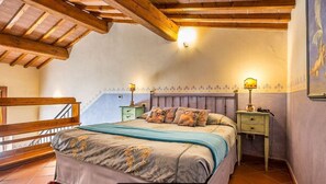 A bedroom with a wooden ceiling, a bed with a floral bedspread, two bedside tables with lamps, and a staircase.