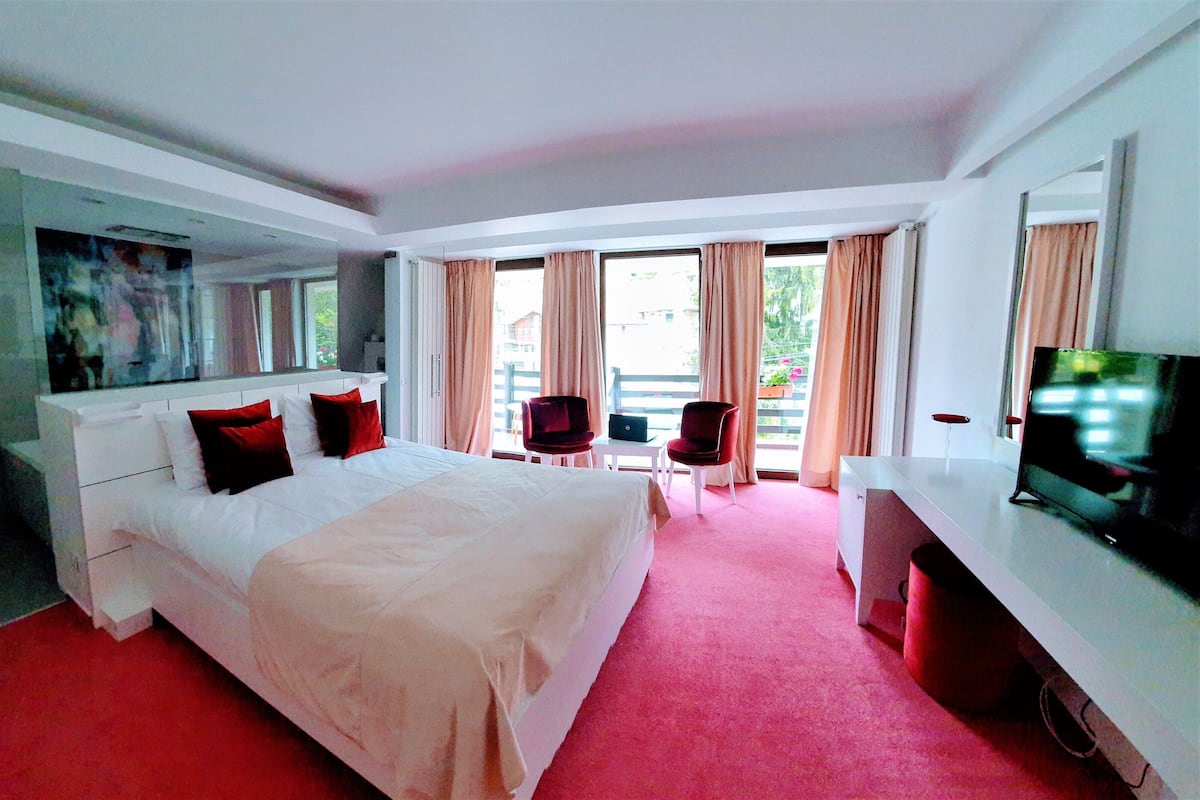 A hotel room with a large bed, a desk, a chair, a TV, and a balcony with curtains.