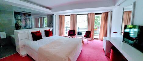 Habitación de hotel con una cama grande, un escritorio, una silla, un televisor y un balcón con cortinas.