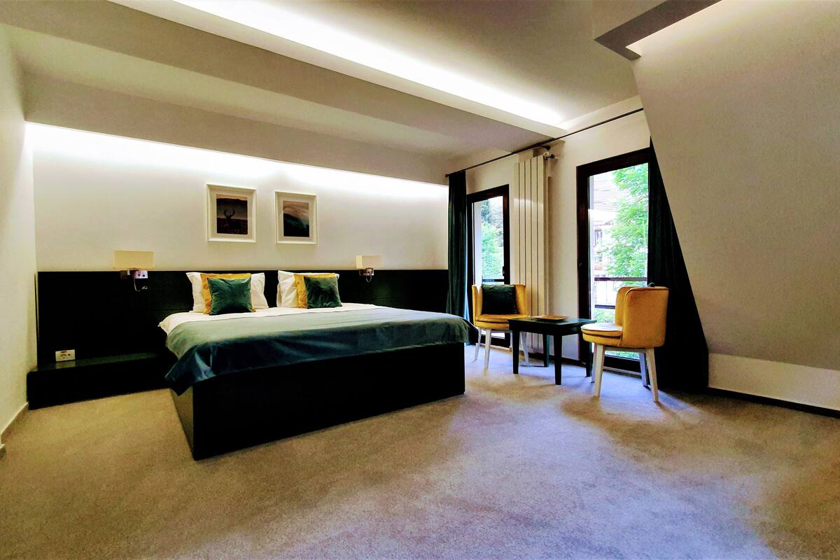 A modern hotel room with a large bed, a desk, and a chair. There are two wall-mounted lamps and a window with a view of greenery.