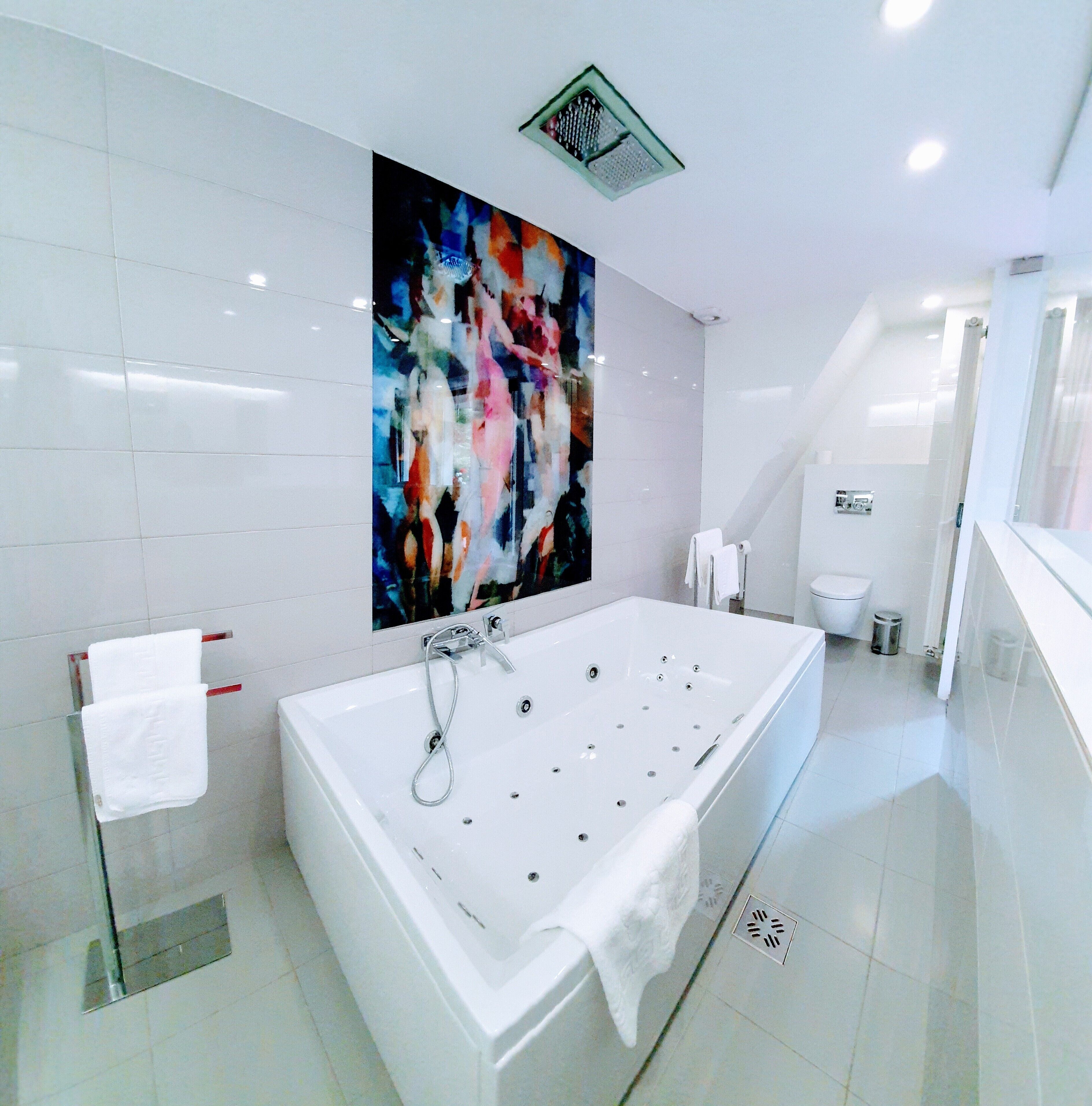 A modern bathroom with a large bathtub, white tiles, a colorful abstract painting, and a visible toilet.