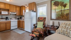 A kitchen with wooden cabinets, a refrigerator, and a microwave. There is a chair with a colorful cushion, a dining table with a chair, and a painting on the wall. - Porter's Cottage....Tiny Home Located At Stillwater Ranch (Laurel Hill)