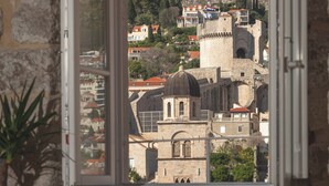 Una habitación con un amplio ventanal con vistas a un pueblo histórico, un televisor sobre un mueble bajo y una silla de madera.