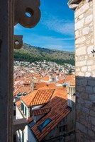 A view from a stone building with a window, overlooking a town with red-tiled roofs and a hill in the background.