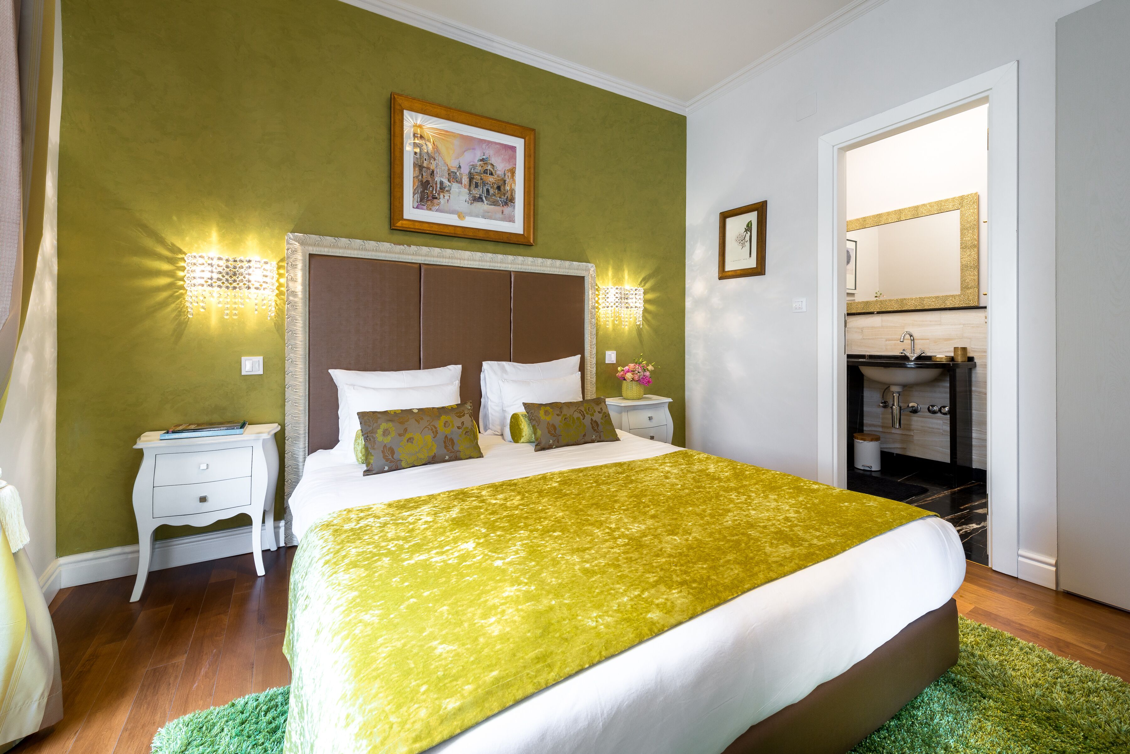 A neatly arranged bedroom with a large bed, bedside tables, a nightstand, a green rug, and a bathroom visible through an open door.