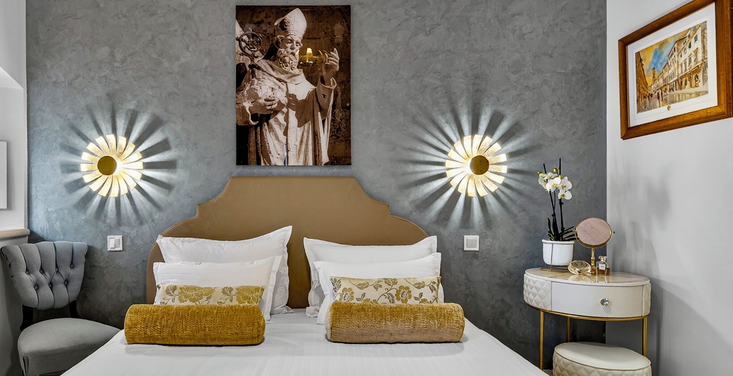 Un dormitorio con una cama grande, dos apliques de pared, una obra de arte enmarcada, una mesita de noche con un espejo y una silla.