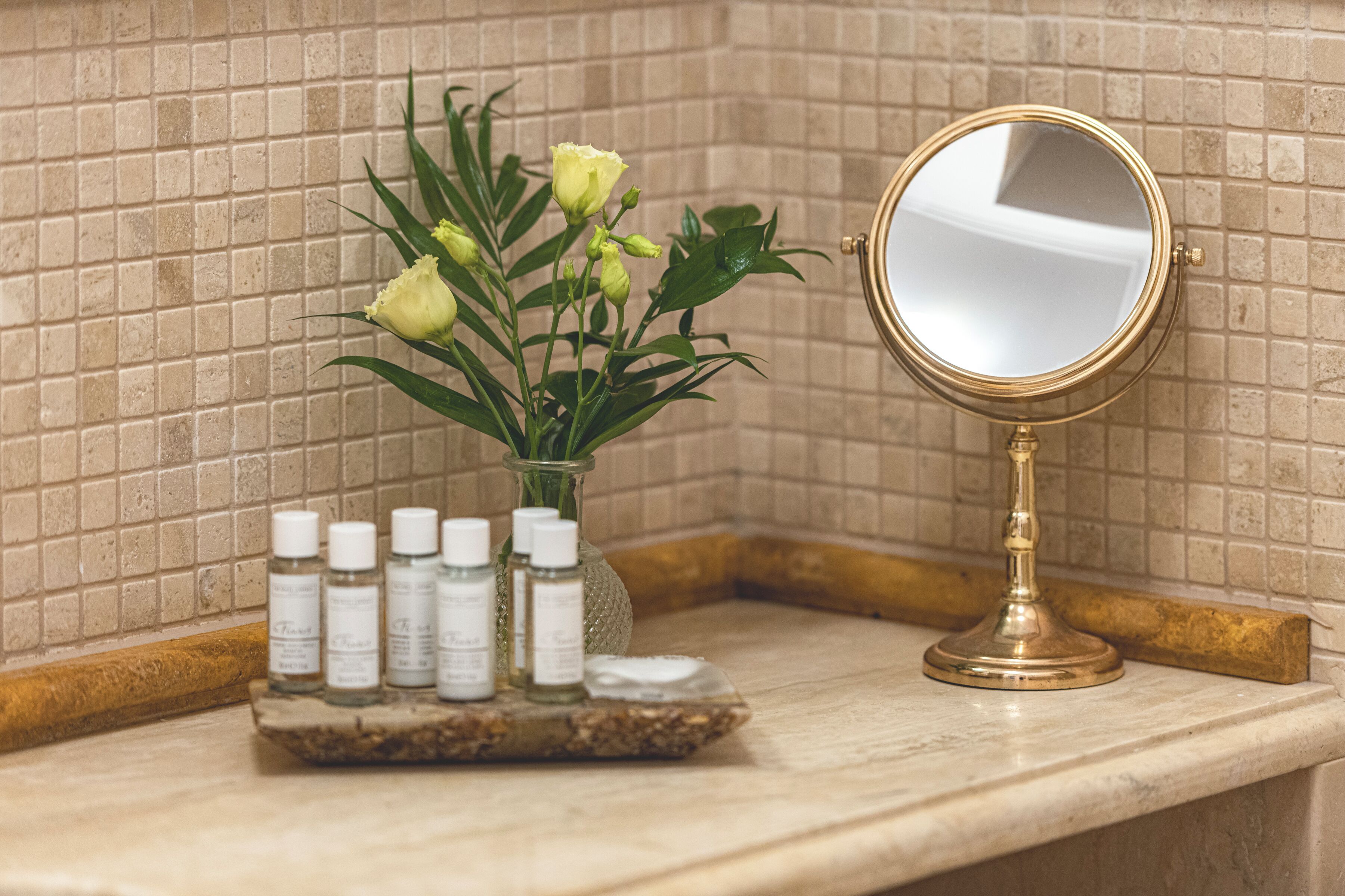 A bathroom vanity with a mirror, a vase with flowers, and several bottles of toiletries.