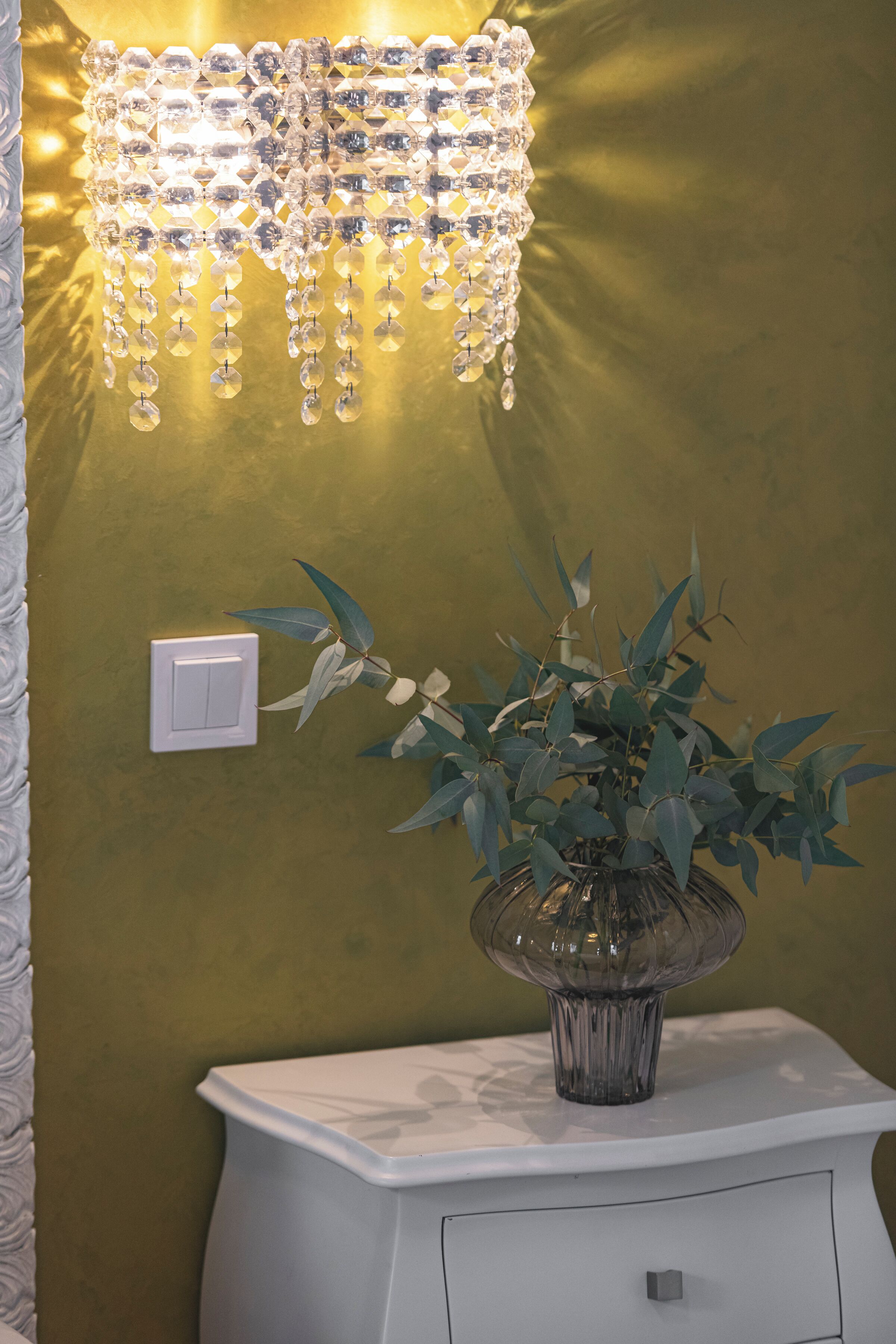 A white bedside table with a vase of green leaves, a wall-mounted light fixture, and a light switch.
