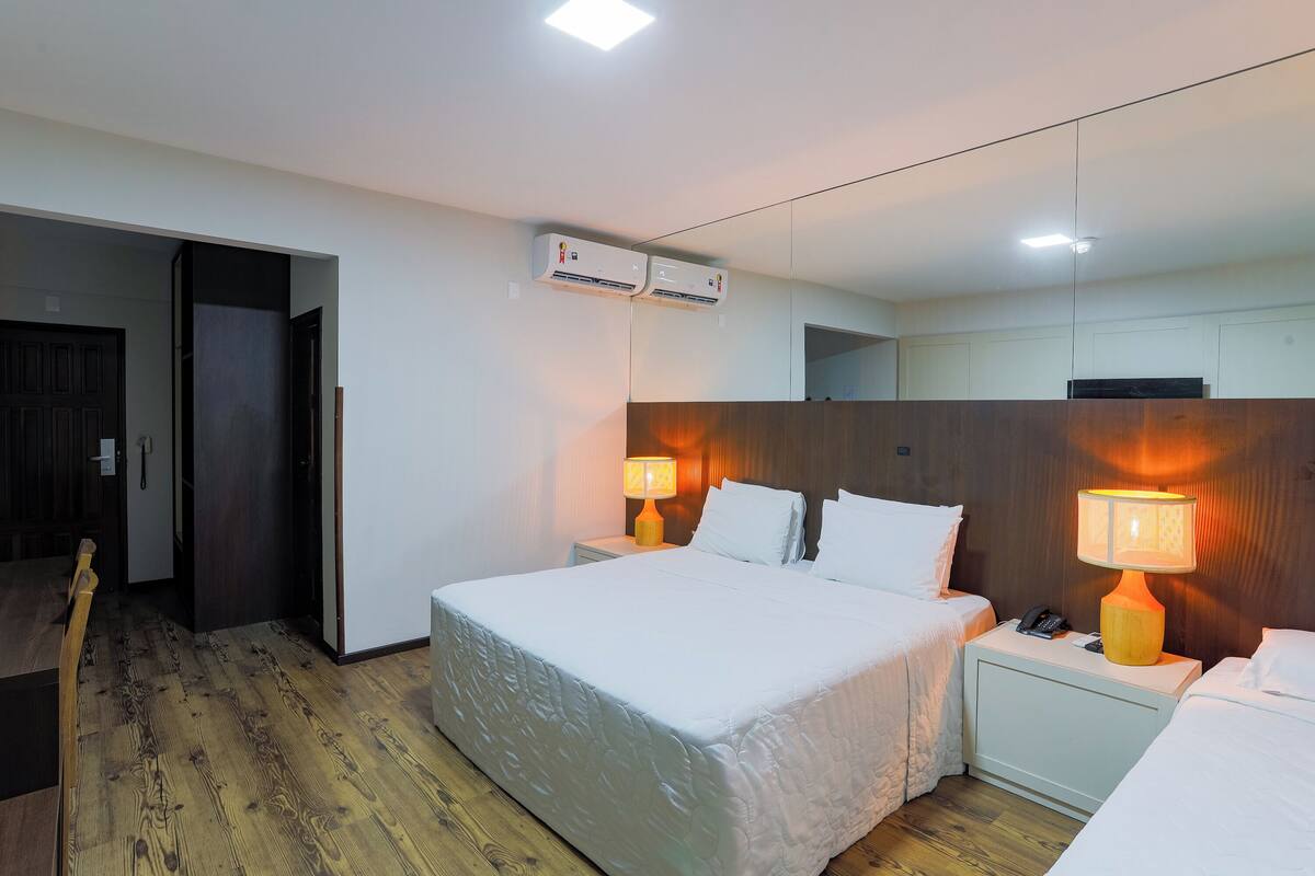 A hotel room with a large bed, two bedside tables with lamps, an air conditioning unit, and a wooden floor.
