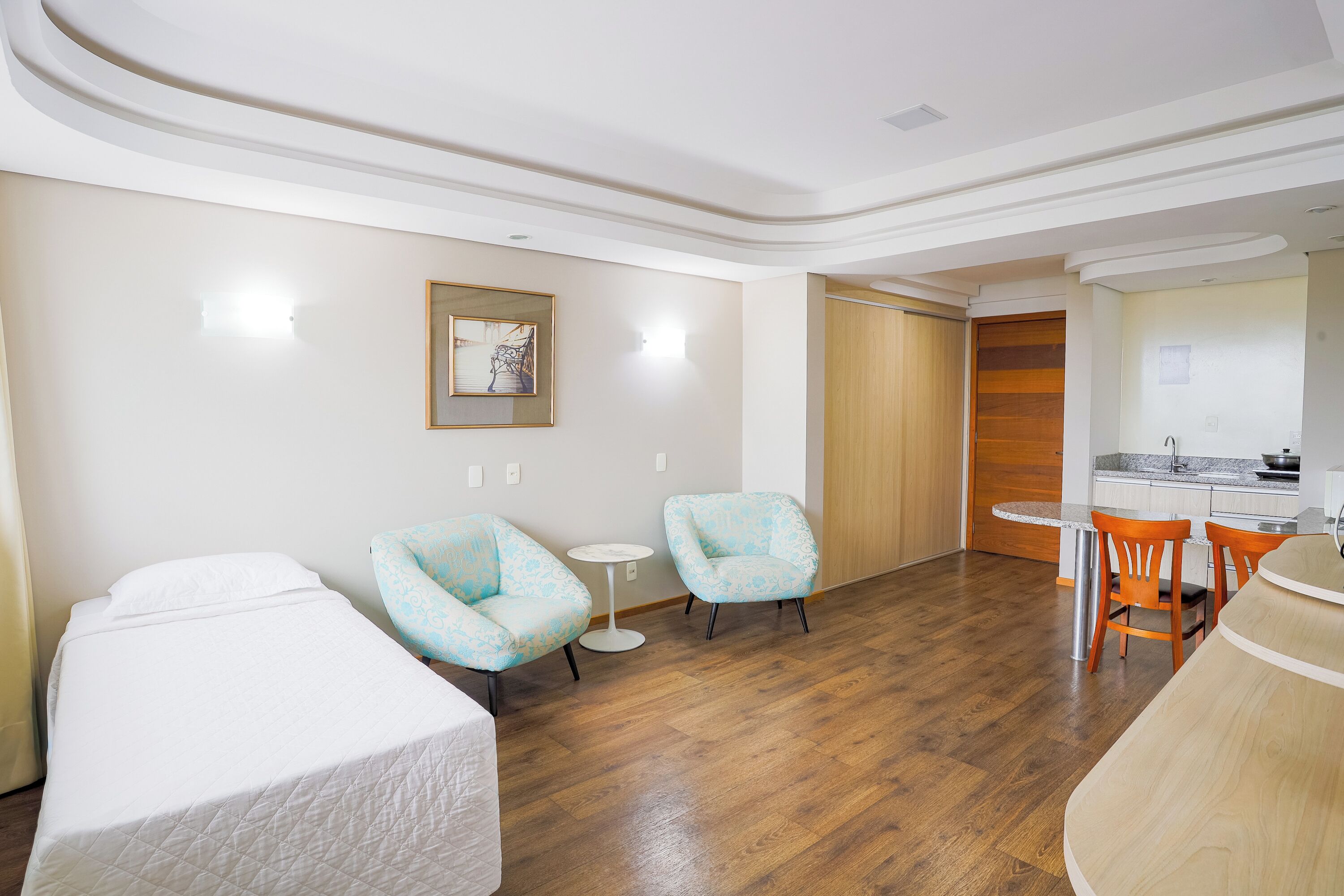 Habitación de hotel con dos camas, una mesita redonda pequeña, dos sillones con estampado y una kitchenette. Se observa también una puerta de madera.