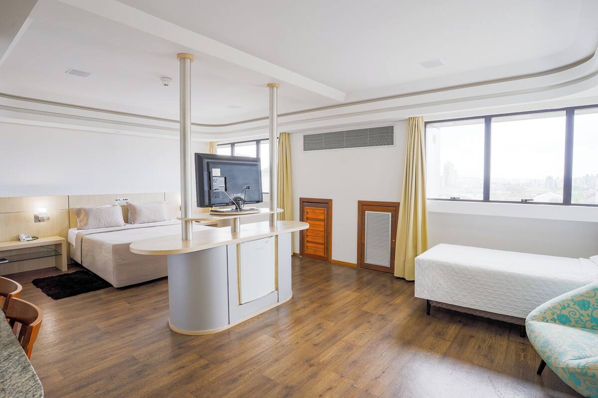 A modern hotel room with a central island, a bed, a sofa, a TV, and a large window with a city view.