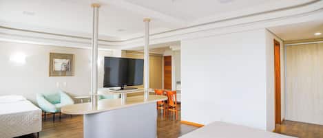 A modern hotel room with a large bed, a small sitting area with a TV, and a separate dining area with orange chairs.