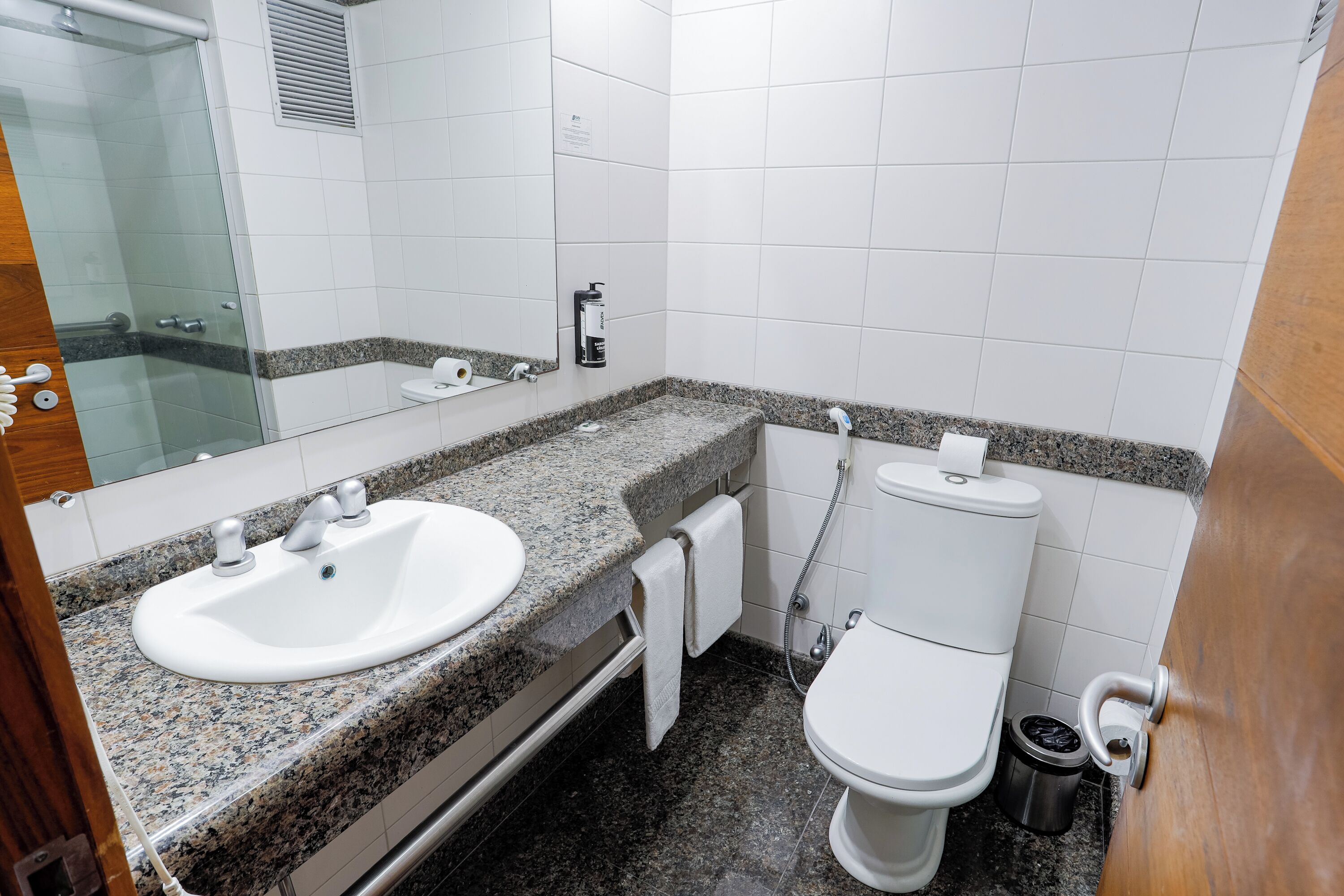 Un baño con lavamanos de encimera de granito, espejo, inodoro y cubo de basura.