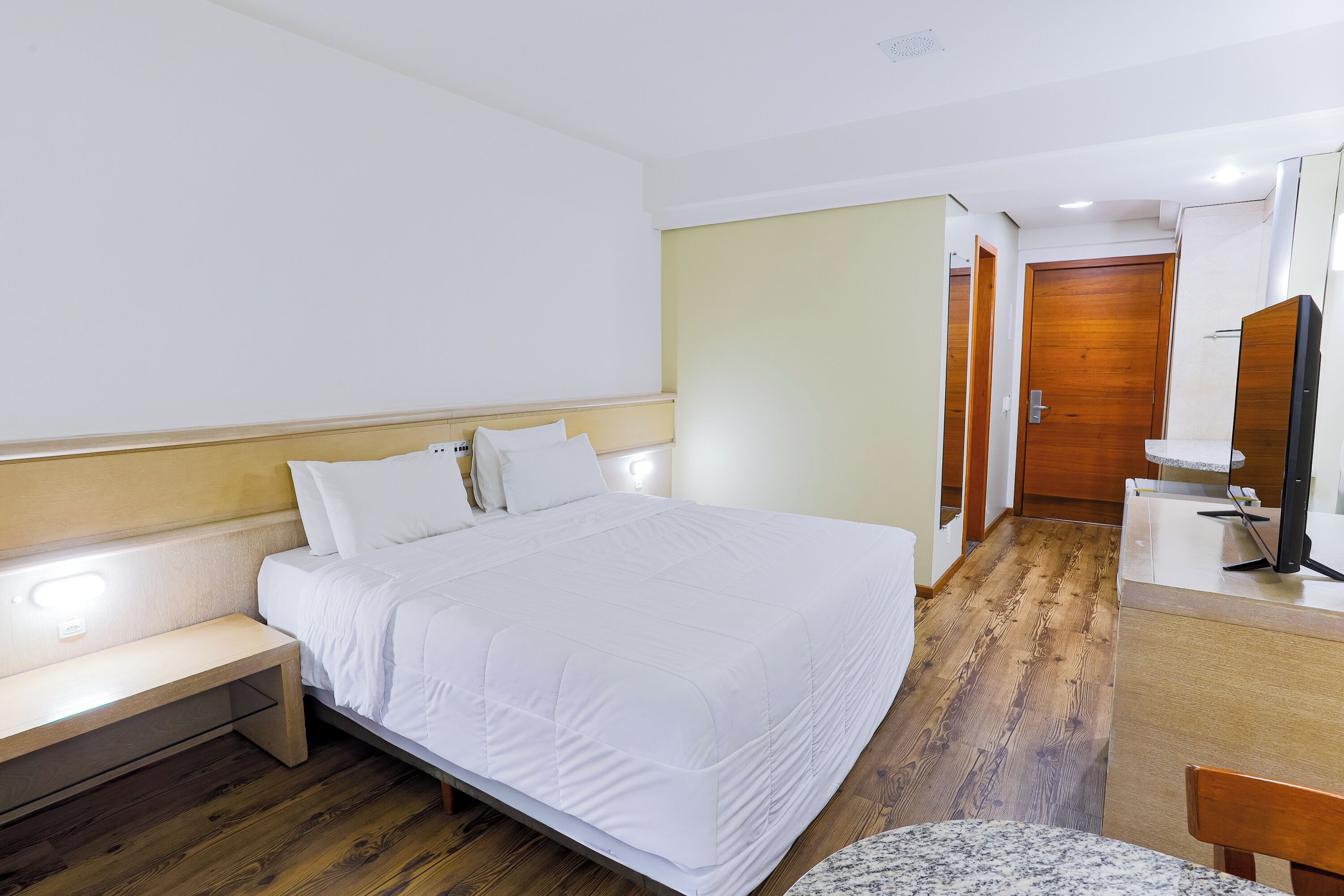 A hotel room with a bed, bedside table, bench, wooden floor, and a small desk with a chair.