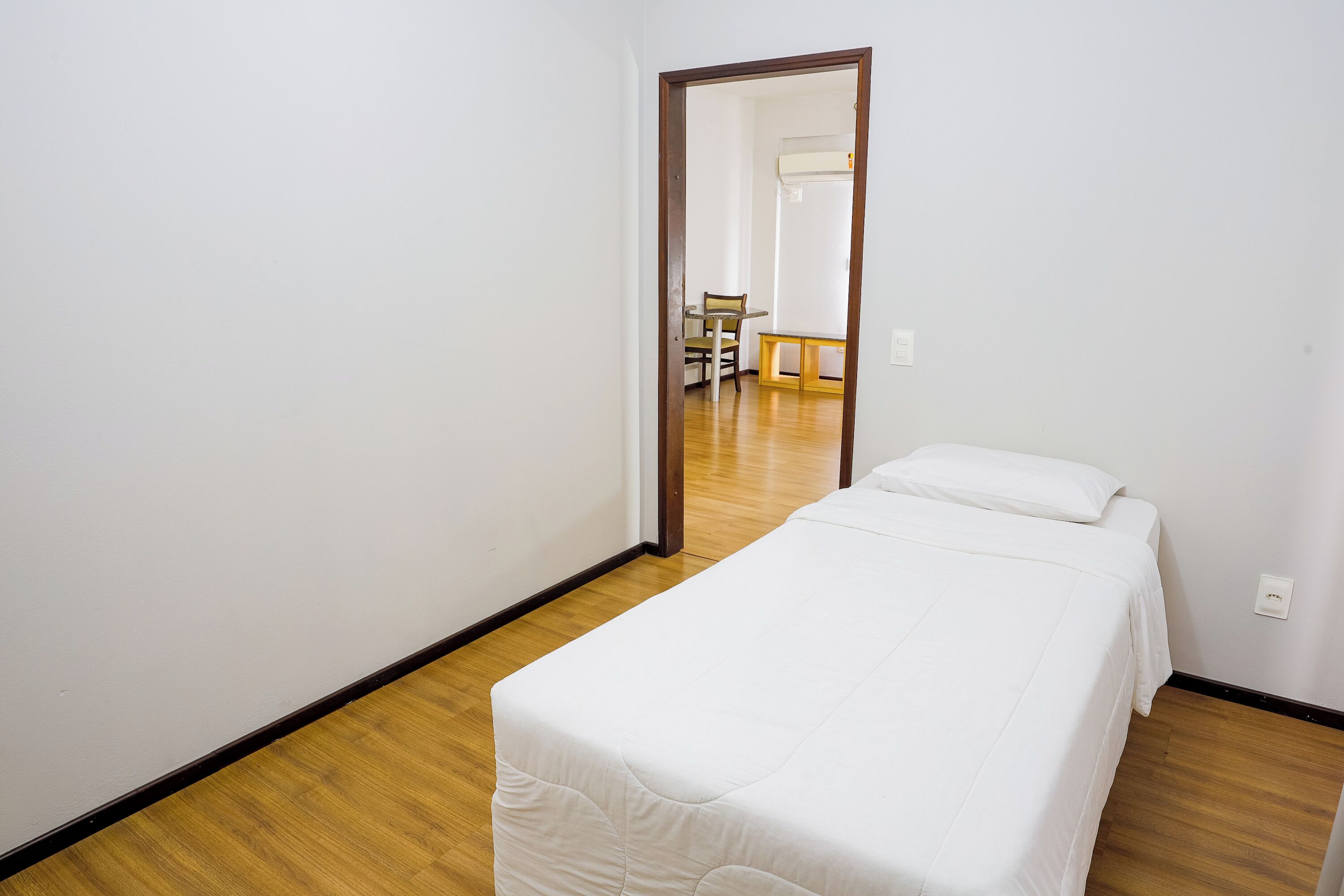 Ein Schlafzimmer mit einem Einzelbett, Holzboden und einem Spiegel, der einen Essbereich mit Tisch und Stühlen reflektiert.